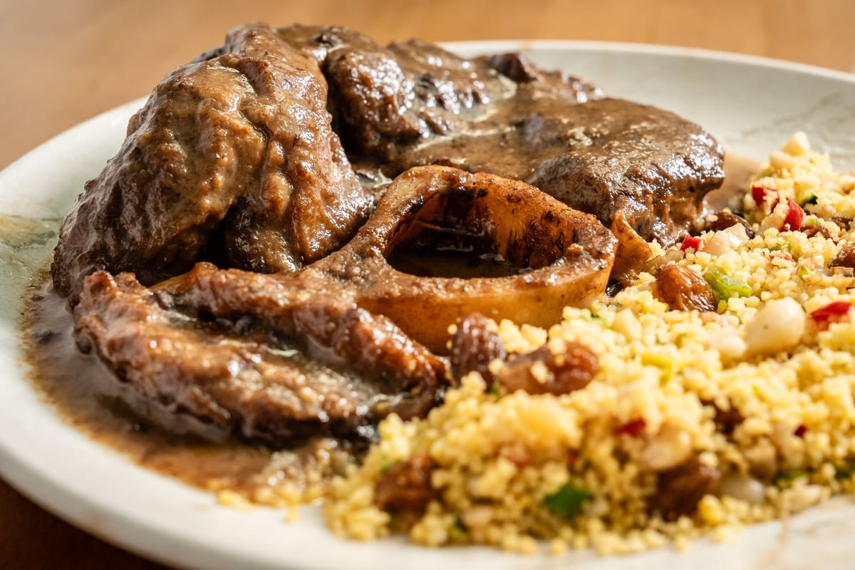 Plato de ossobuco de ternera cocinado en salsa oscura, servido sobre un lecho de risotto amarillo con trozos de verduras. La